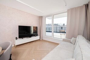 Un salon moderne doté d’un téléviseur à écran plat, d’un canapé, d’un fauteuil et d’une grande fenêtre donnant sur la ville.