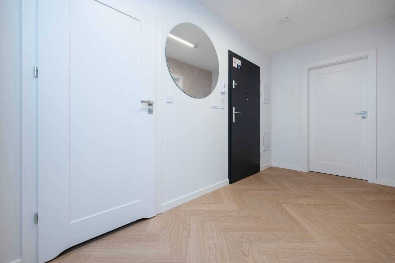 Um corredor com portas brancas, uma porta preta com um espelho circular e piso de madeira.