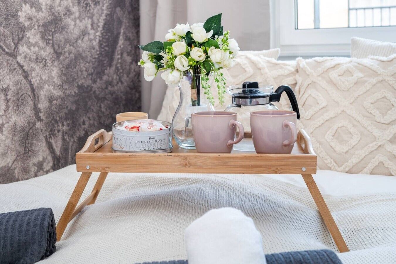 Un plateau avec une théière, deux tasses, un vase de fleurs et une boîte de biscuits, le tout posé sur un lit agrémenté d’oreillers et d’une couverture.