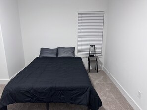 Ein Schlafzimmer mit einem Bett, zwei Kissen, einem Nachttisch und einem Fenster mit Jalousien.