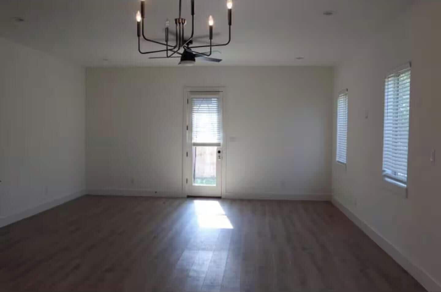 A spacious room with a wooden floor, a chandelier, a door with a window, and a wall-mounted light fixture.