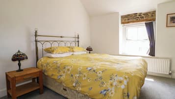 Quarto com cama, mesa de cabeceira, abajur, janela com cortinas e radiador.