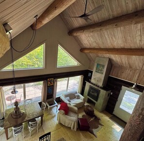 Un interior rústico con techo alto, vigas de madera, zona de comedor con mesa y sillas, chimenea y ventanas amplias que dejan entrar luz natural.