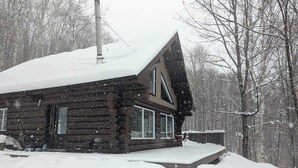 Une cabane en rondins, dont le toit est recouvert de neige et qui possède une cheminée, est entourée d’un paysage enneigé.