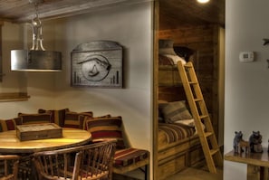 Um quarto aconchegante com uma cama beliche de madeira, uma mesa de jantar com cadeiras, um candeeiro pendente e um relógio de parede.