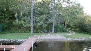 A wooden dock extending over a calm body of water, surrounded by trees and a grassy area with outdoor seating.