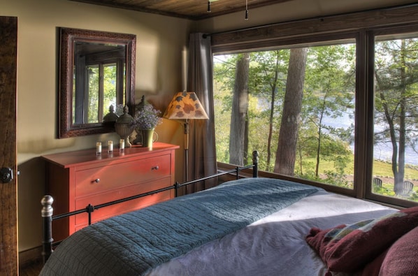 A bedroom with a bed, dresser, mirror, lamp, and a view of trees and water.