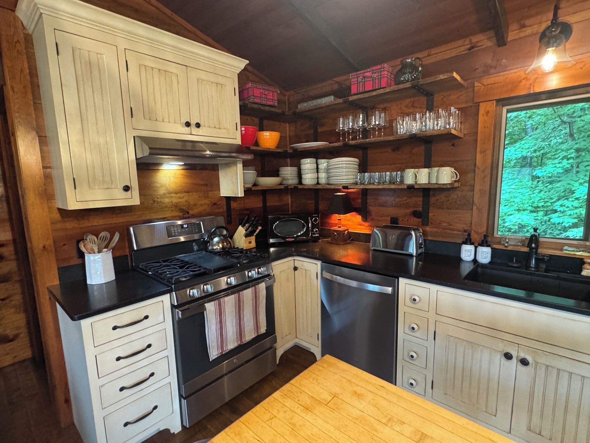 A kitchen with wooden cabinets, a stove, a dishwasher, and a window with a view of trees.