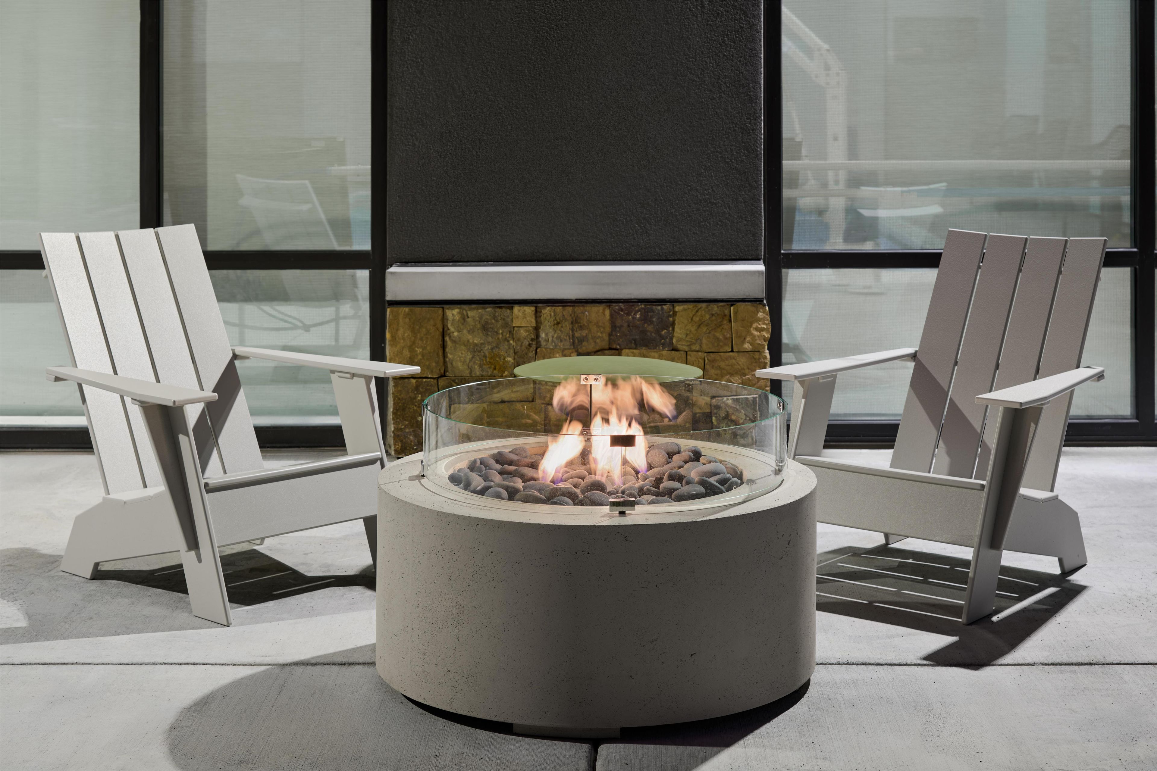 A fire pit with two Adirondack chairs, a stone fireplace, and a view through large windows.