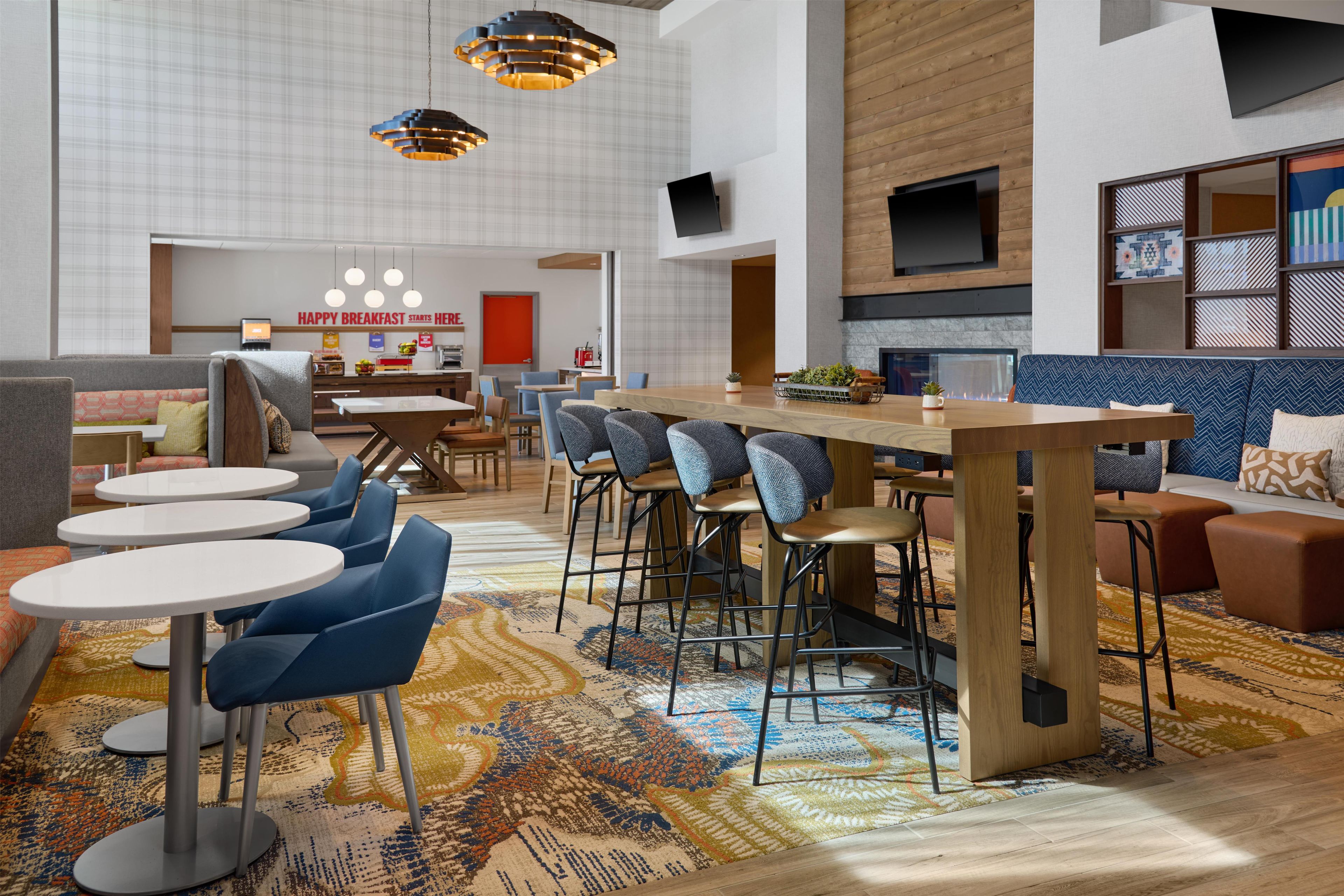 A modern hotel lobby with a central bar, seating areas, and a breakfast counter.