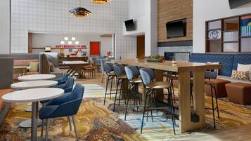 A modern hotel lobby with a central bar, seating areas, and a breakfast counter.