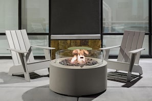 A fire pit with two Adirondack chairs, a stone fireplace, and a view through large windows.