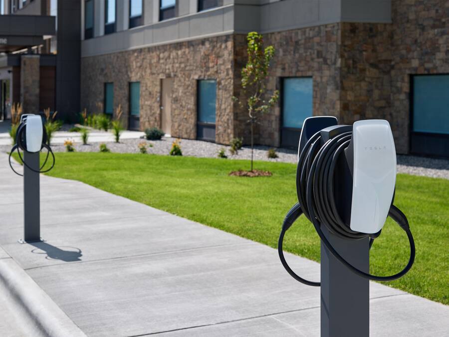 Una fila de estaciones de carga para vehículos eléctricos a lo largo de un camino de hormigón, con un edificio de piedra y cristal al fondo.