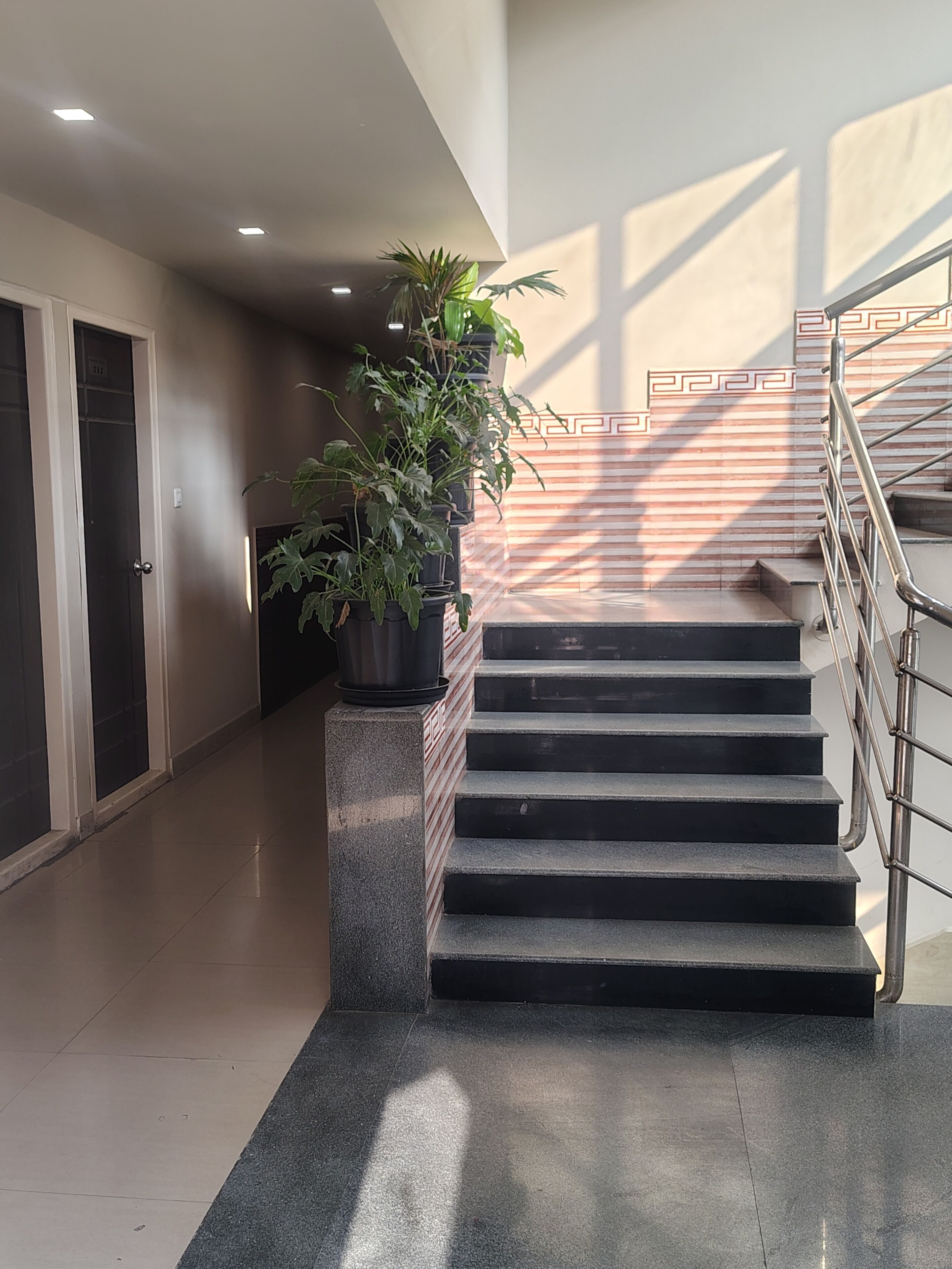 Una escalera con barandilla de metal que conduce a un piso superior, una planta en maceta sobre un pedestal, y una puerta a la izquierda.