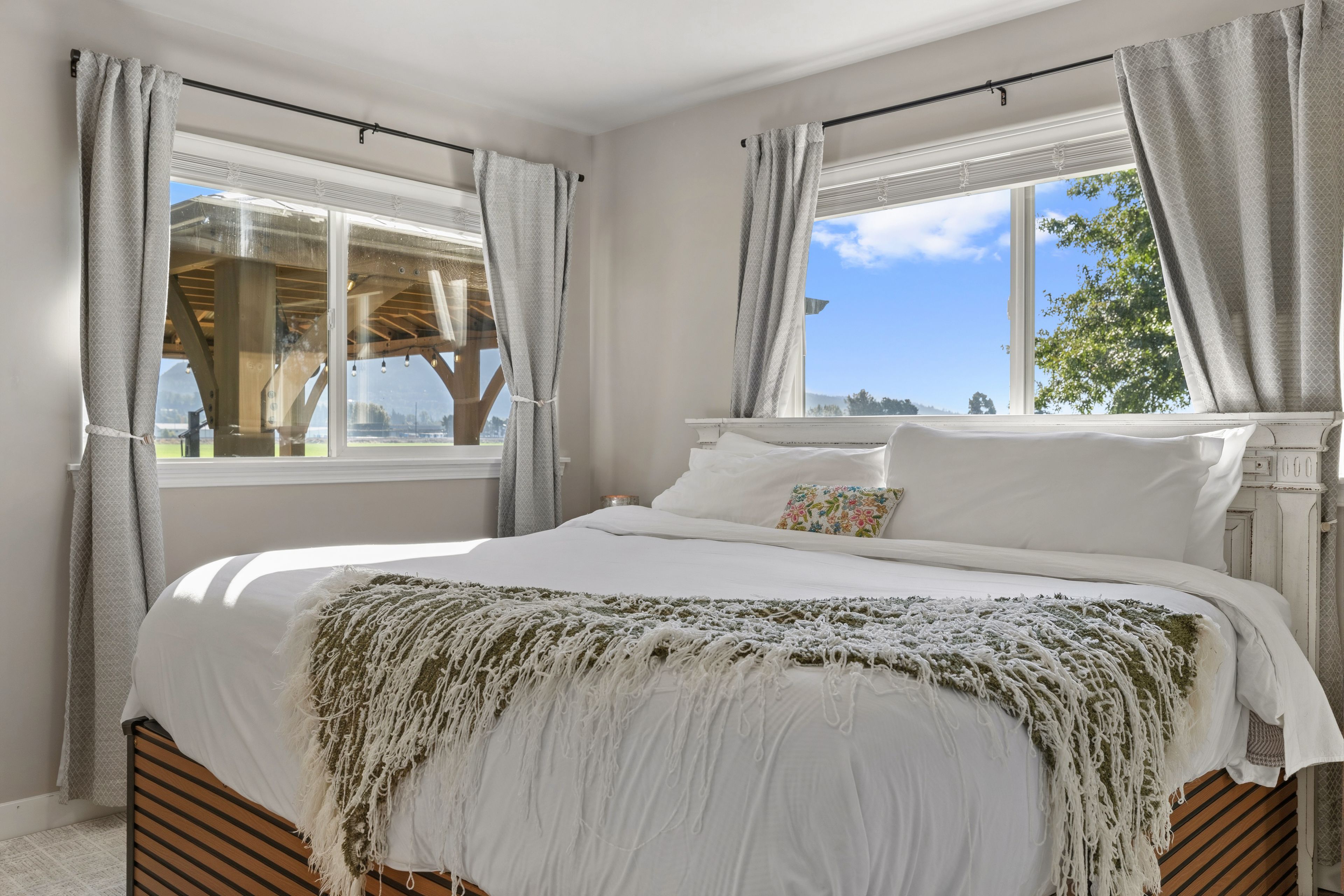 A bedroom with a large bed, two windows with curtains, a wooden headboard, and a view of the outdoors.