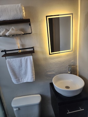 Un baño con un lavamanos blanco, un espejo con luz y dos toallas en un perchero.