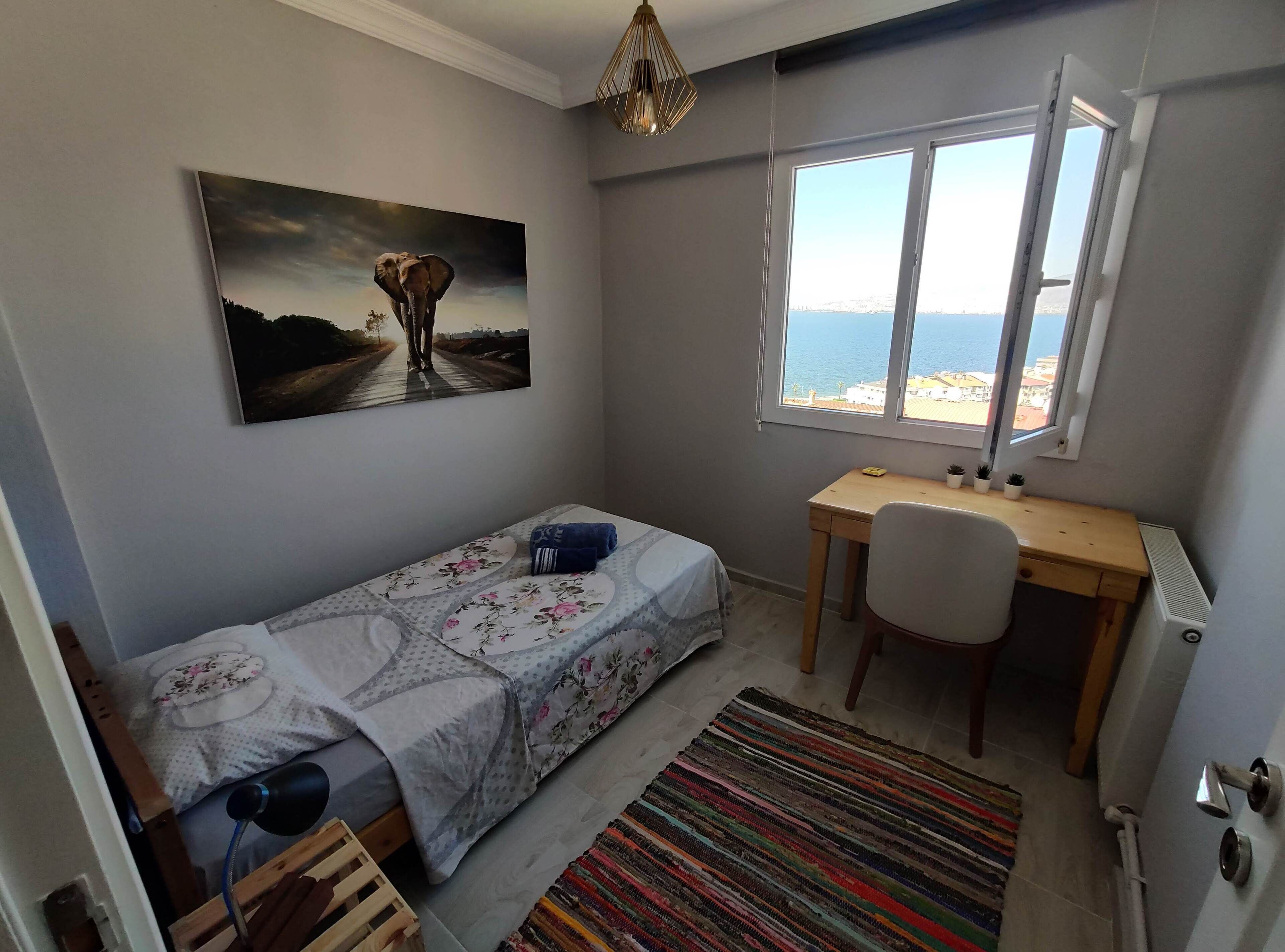 A small, neatly arranged room with a bed, a desk, a chair, a radiator, and a window with a view of the sea.