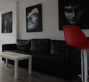 Ein modernes Wohnzimmer mit einem schwarzen Ledersofa, einem weißen Couchtisch und einem roten Sessel. An der Wand hängen drei gerahmte Kunstwerke.
