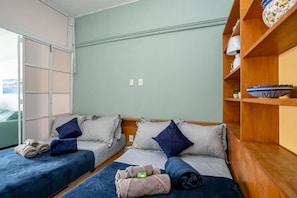 Una habitación con dos camas, cada una con colcha y almohadas azules, un estante de madera con objetos decorativos y una puerta que da a otra habitación.