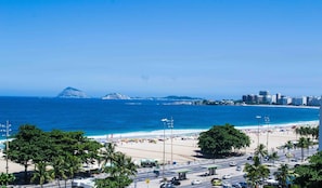 A beach with a clear view of the ocean, a city skyline in the background, and a road with vehicles and palm trees in the foreground. - Tio Sam  (Rio de Janeiro)