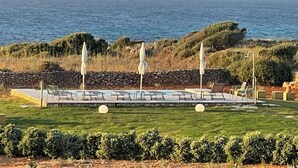 Zona junto a la piscina con sillones de descanso y sombrillas, una pared de piedra y vistas al mar y acantilados.