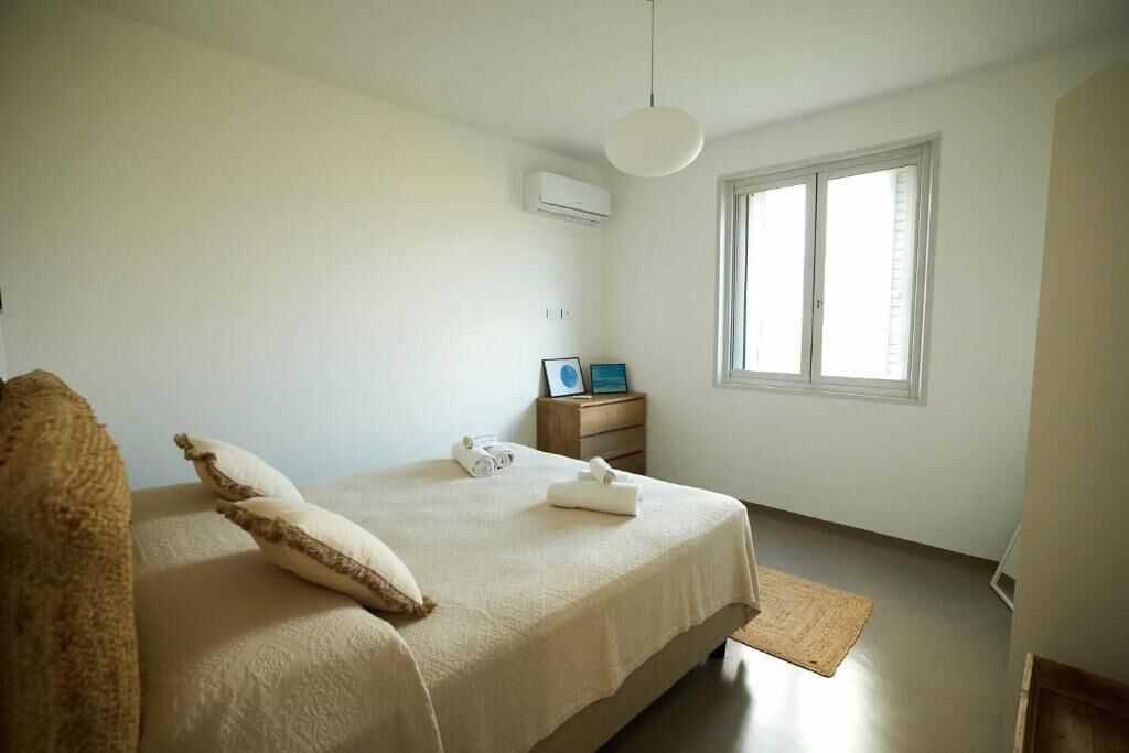 A bedroom with a bed, a bedside table, a window, and an air conditioning unit.