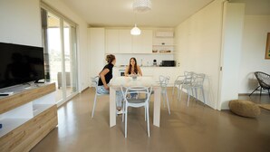 A modern kitchen with white cabinets, a dining area with a table and chairs, and a television on a wooden stand. - Villa Zabbara (Favignana)