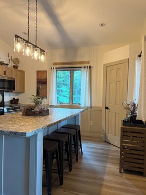 A kitchen with a large island, bar stools, a hanging light fixture, and a window with curtains.