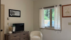 Un dormitorio con cama, una silla, un televisor sobre un aparador de madera, un ventilador de techo y una ventana con cortinas.