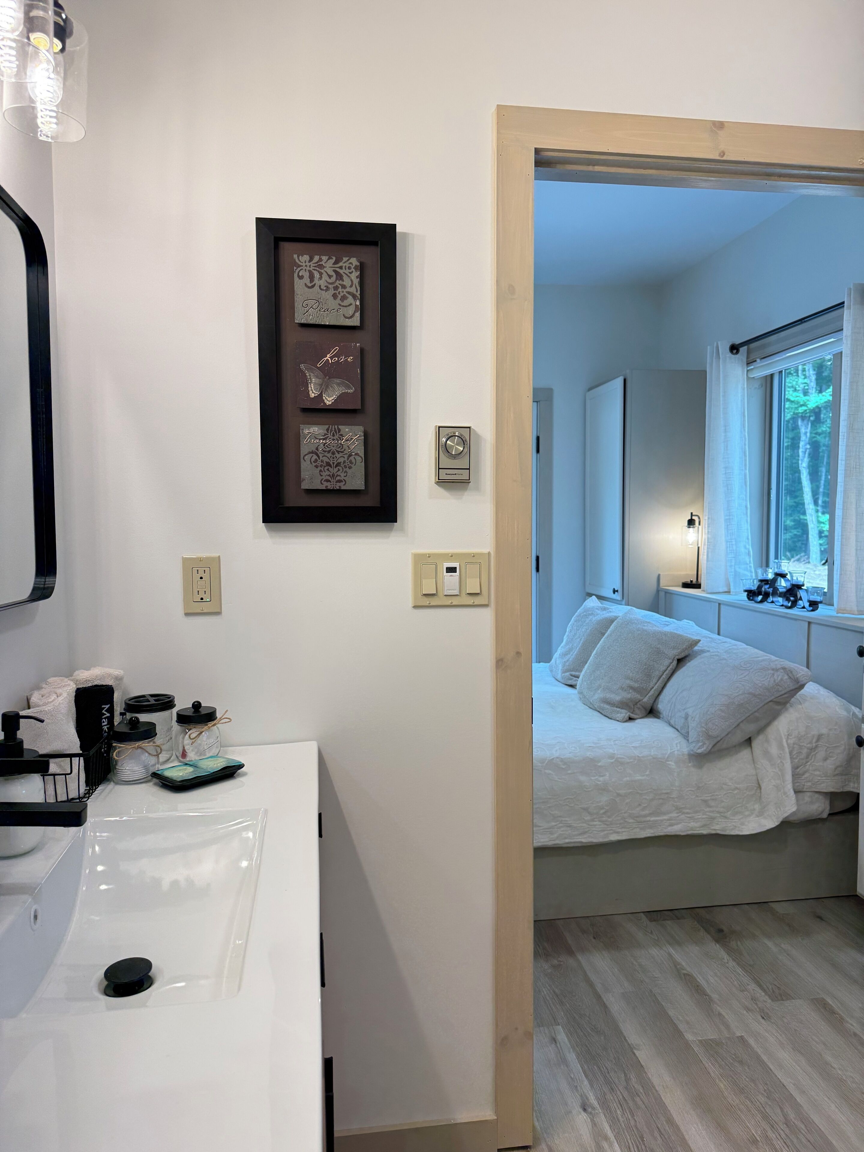 A hotel room with a white bed, a window with a view of trees, a bathroom with a white sink and a mirror, and a wall with framed artwork.