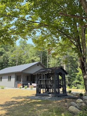 A house with a covered outdoor seating area, surrounded by trees and a gravel driveway.