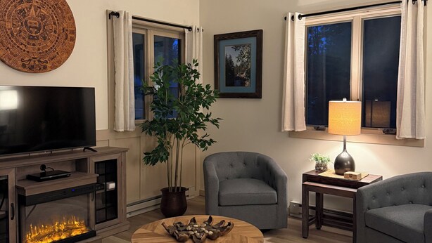Una sala de estar con una televisión de pantalla plana, un ventilador de techo, una chimenea, una mesa de centro redonda y dos sillones grises.