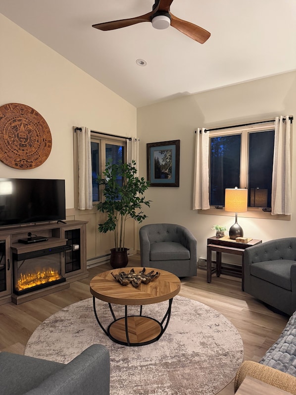 A living room with a flat-screen TV, a ceiling fan, a fireplace, a round coffee table, and two grey armchairs.