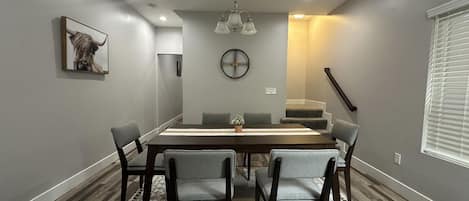 A dining area with a rectangular table, six chairs, a clock on the wall, and a staircase leading to an upper level.
