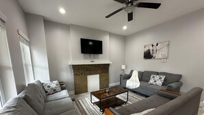 A modern living room with a fireplace, grey sofas, a coffee table, and a ceiling fan. - 2BR Sunny Haven Near Short North (Columbus)