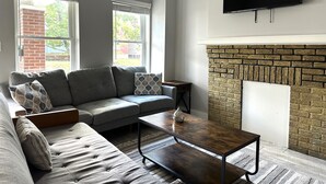 A living room with a grey sofa, a coffee table, a fireplace, and a flat-screen TV mounted on the wall. - 2BR Sunny Haven Near Short North (Columbus)