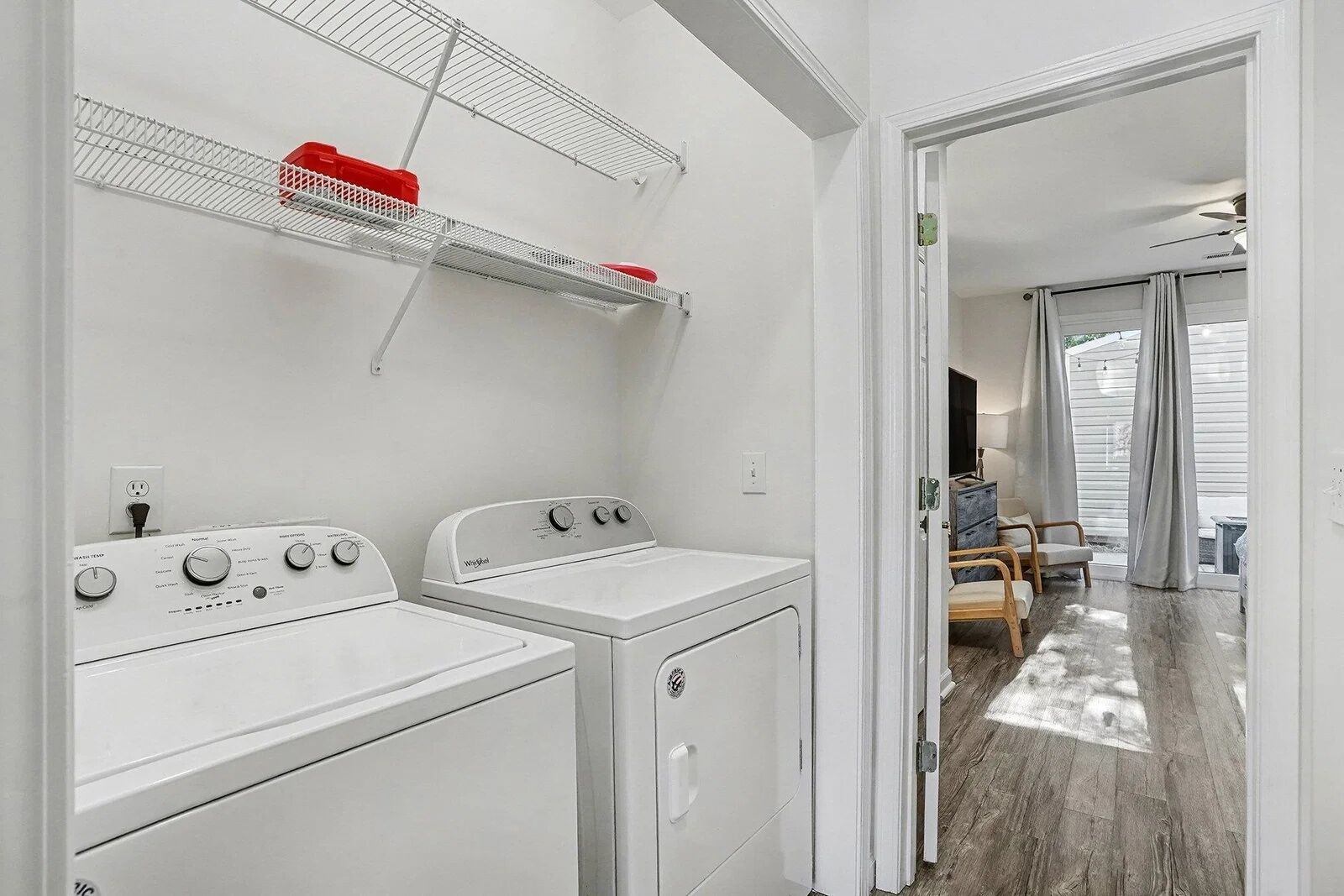 A laundry room with two white washing machines, a metal drying rack, and a door leading to a living area with a sofa and a TV.