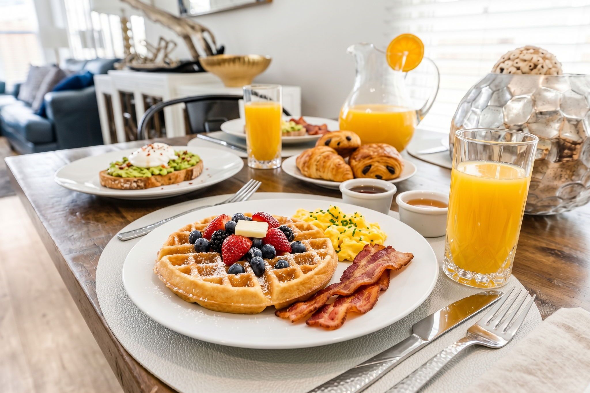 Une table dressée pour le petit-déjeuner avec des gaufres, du bacon et des fruits sur une assiette, une carafe de jus d’orange et un verre de jus d’orange.