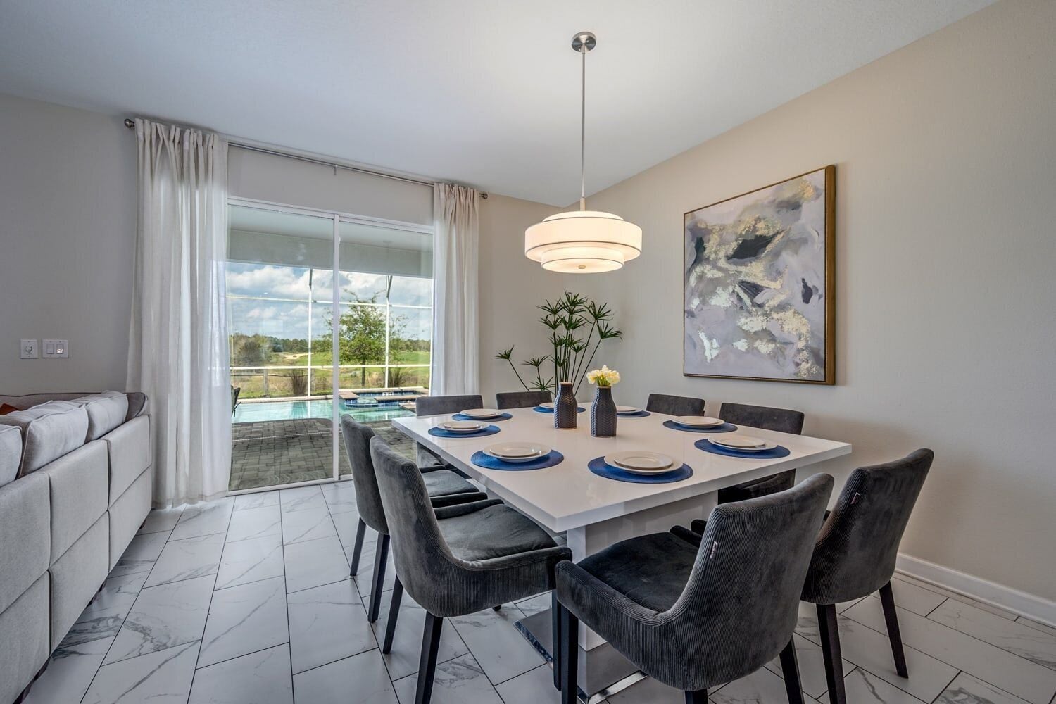 A dining area with a white table, dark chairs, a hanging light fixture, and a painting on the wall.