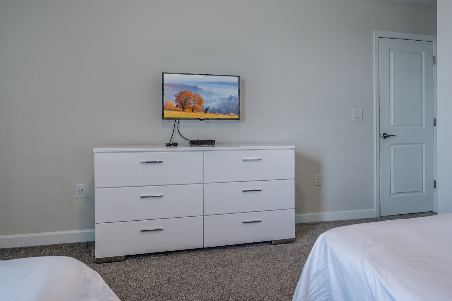 Une chambre avec une commode blanche, un téléviseur fixé au-dessus, et une porte sur le côté droit.