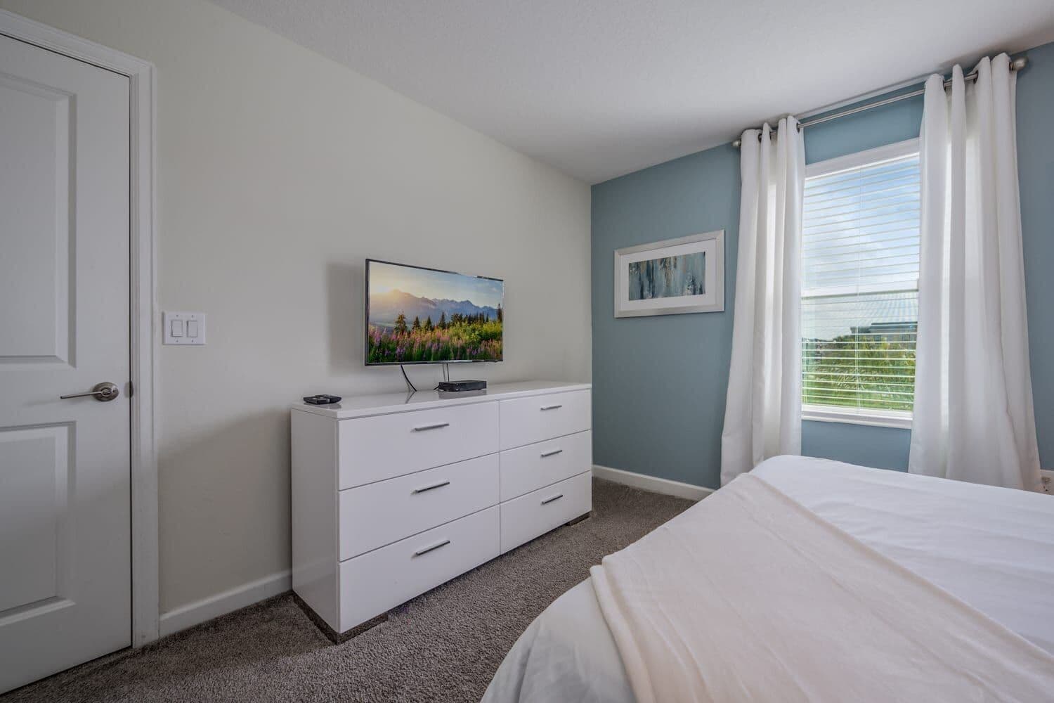 A bedroom with a bed, a dresser, a television on the wall, and a window with curtains.