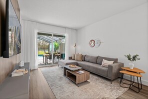 A modern living room with a grey sofa, a coffee table, and a TV mounted on the wall. - Modern 5BR Home Near Disney With Private Pool (Davenport)