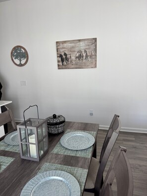 A dining area with a wooden table, two chairs, a candle holder, a decorative plate, and a wall-mounted painting. - Cozy 2 bedroom 2 bath villa in exciting new Eastport.  Village of Edenfield. (Sumterville)