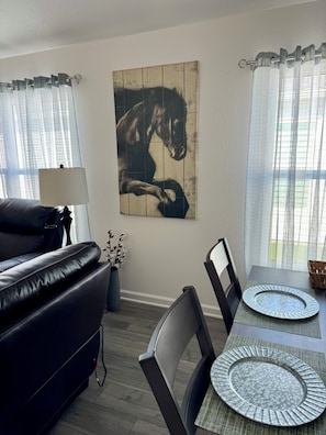 A dining area with a table set for two, a black leather sofa, a wall-mounted painting, and sheer curtains. - Cozy 2 bedroom 2 bath villa in exciting new Eastport.  Village of Edenfield. (Sumterville)