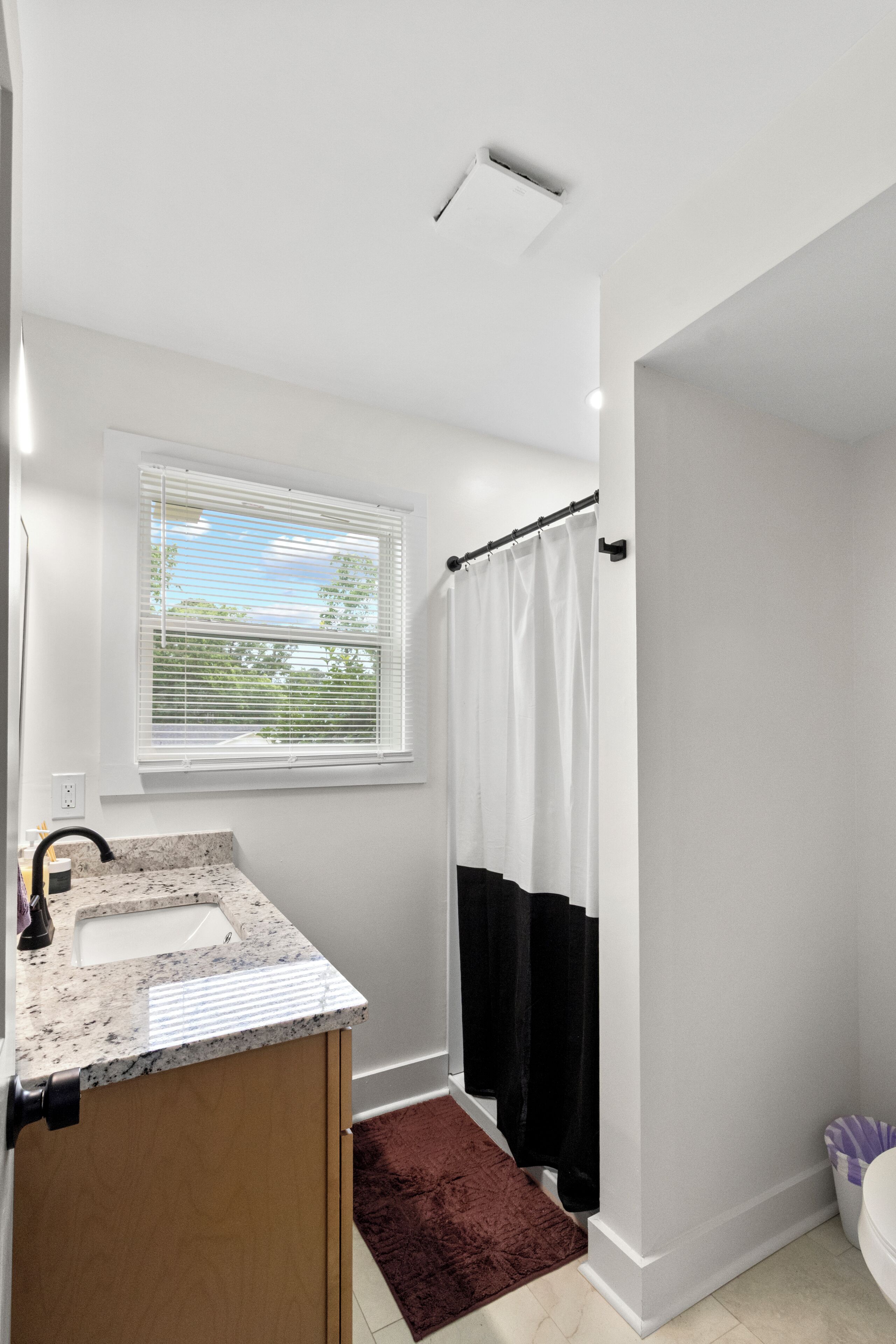A bathroom with a white sink, a window with blinds, a shower curtain, and a marble countertop.