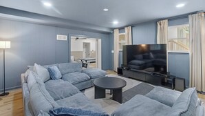 A modern living room with a sofa set, a coffee table, a flat-screen TV, and a dining area in the background.