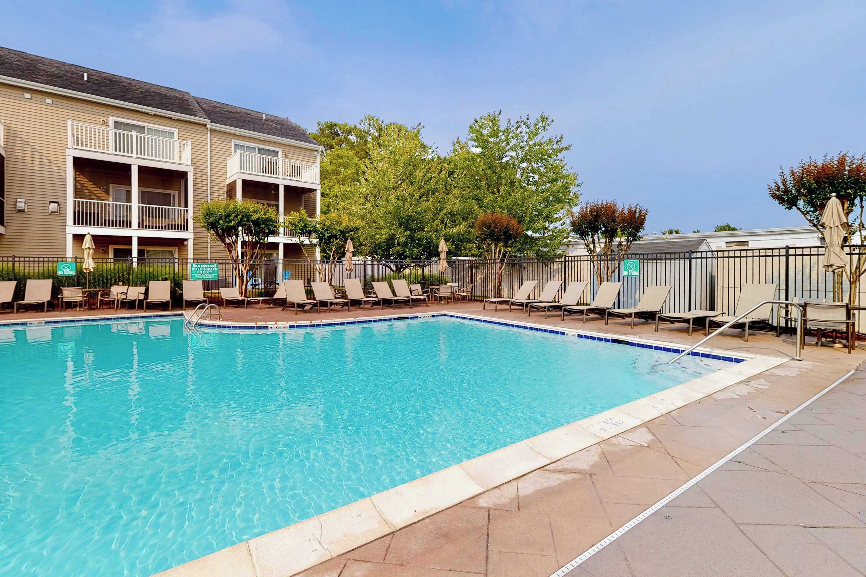 Une piscine dotée de chaises longues et d’une zone délimitée par une clôture, entourée d’immeubles et d’arbres.