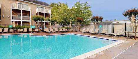 A swimming pool with lounge chairs and a fenced area, surrounded by buildings and trees.