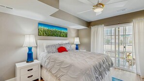 A bedroom with a bed, two bedside tables, a ceiling fan, a window with curtains, and a painting on the wall. - Rehoboth Crossing - 36983 Grindstone Ln (Rehoboth Beach)