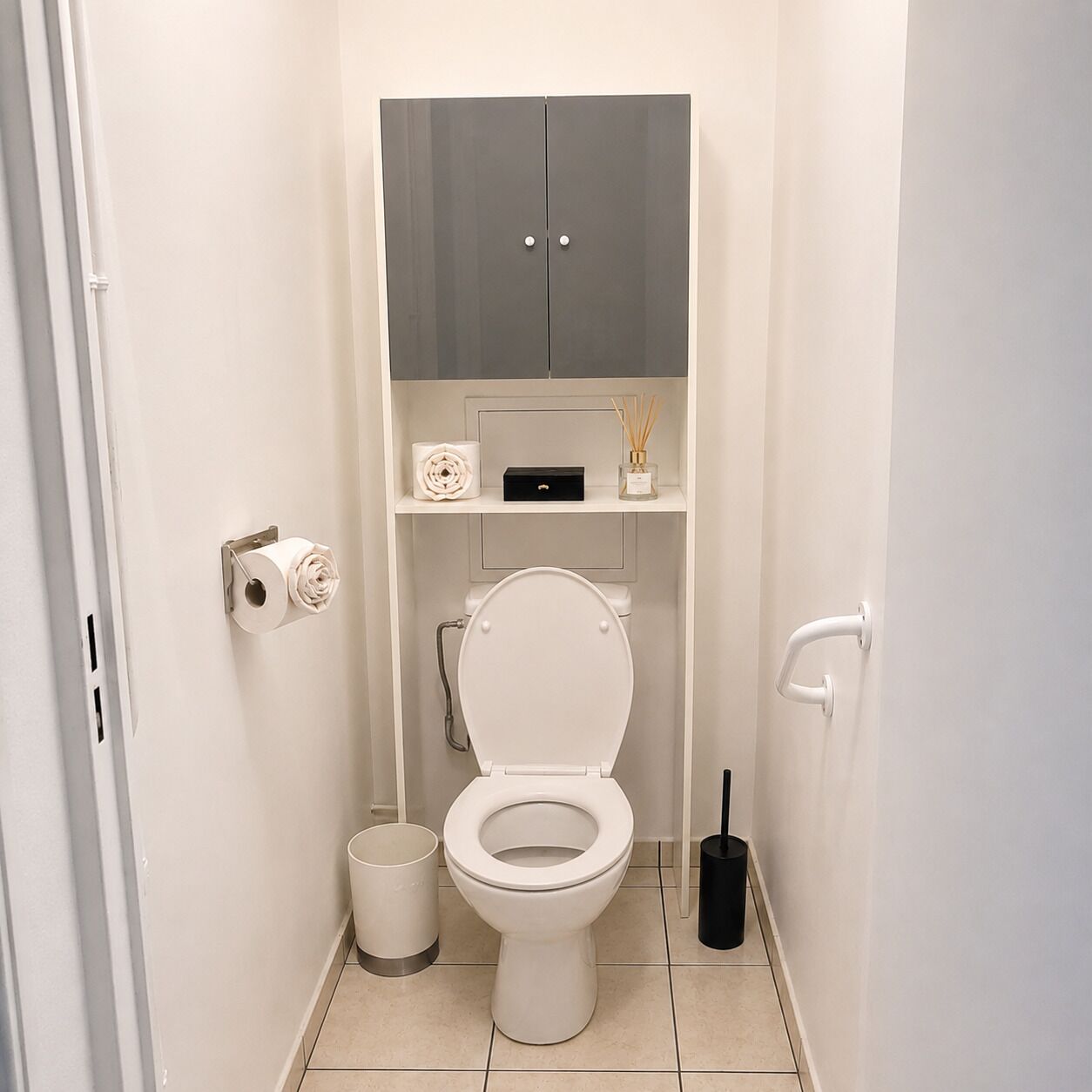 A small, private bathroom with a toilet, a toilet paper holder, a trash bin, a bidet, and a cabinet above the toilet.